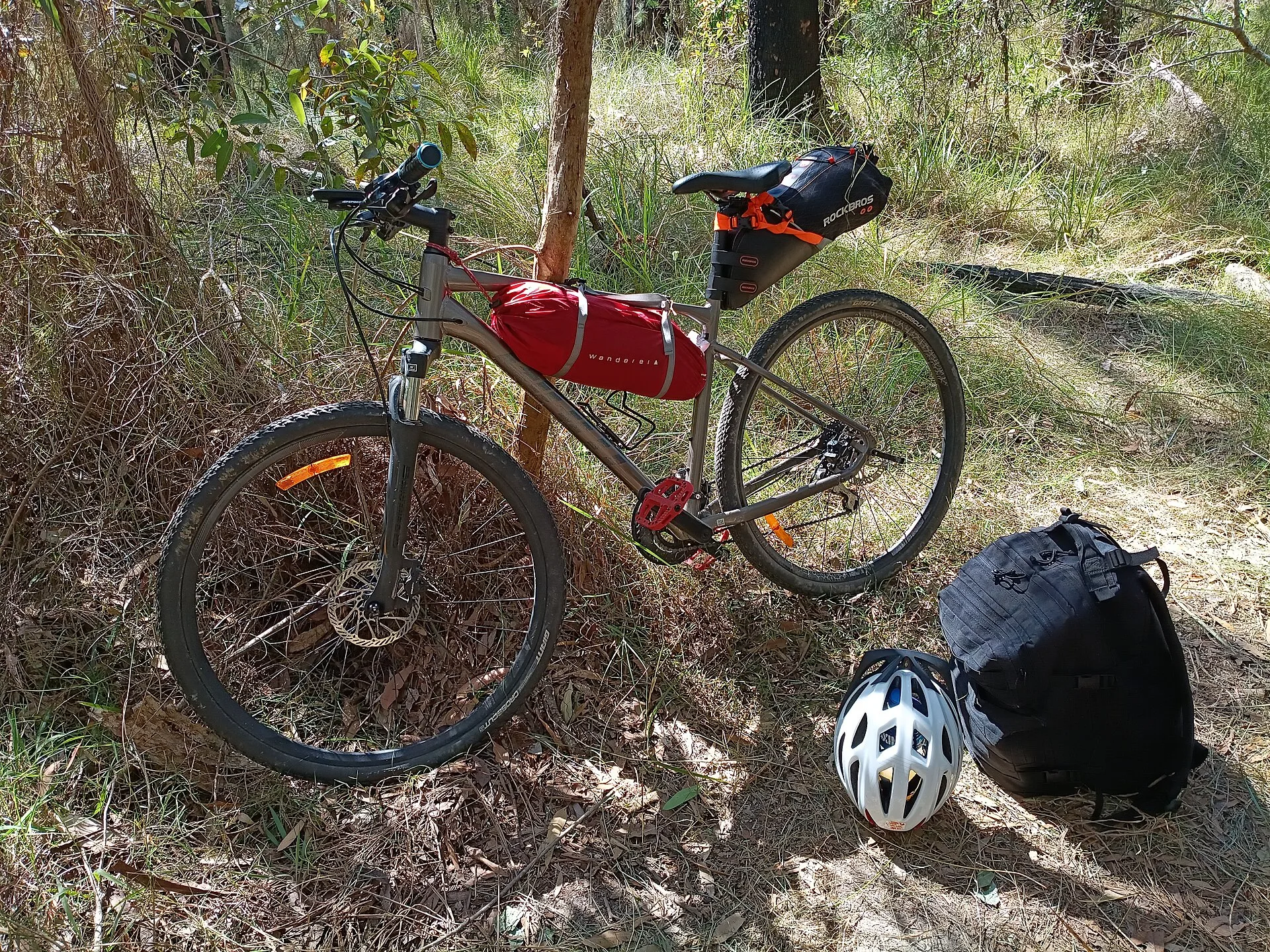 A hybrid bicycle set up for lightweight cycle touring, with a lightweight tent secured beneath the frame and a large waterproof saddle bag carrying clothing, socks and a towel. A MOLLE backpack carries a sleeping bag, compact inflatable mattress, electronics, power bank, toiletries, basic first-aid. Bike itinerary building: book the scarcest item first