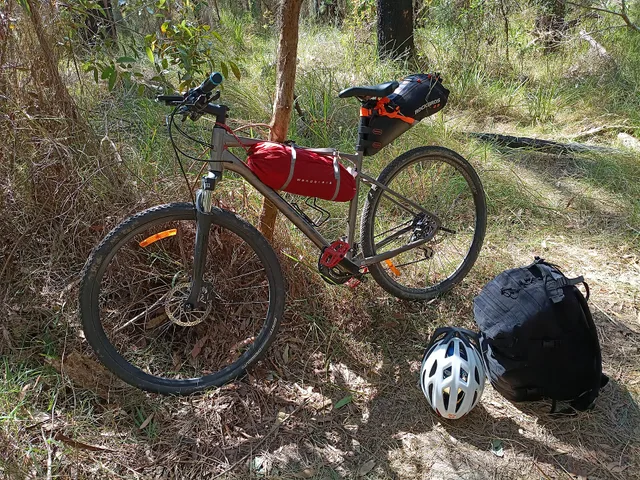 A hybrid bicycle set up for lightweight cycle touring, with a lightweight tent secured beneath the frame and a large waterproof saddle bag carrying clothing, socks and a towel. A MOLLE backpack carries a sleeping bag, compact inflatable mattress, electronics, power bank, toiletries, basic first-aid. Bike itinerary building: book the scarcest item first